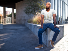 Load image into Gallery viewer, Black man sitting on cement brick wearing white unisex vneck tshirt with Lotus Noir logo in center