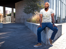 Load image into Gallery viewer, Black man sitting on cement brick wearing white unisex vneck tshirt with Lotus Noir logo in center
